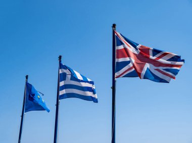 Yunanistan, İngiltere ve AB işaret simgesi. İngiltere, Yunanistan ve Avrupa Birliği bayrak direkleri rüzgarda dalgalanırken bayrak sallıyor. Açık mavi gökyüzü, güneşli bahar günü.