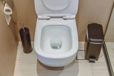 Tuvalet içi, seramik beyaz klozet kapağı açık, siyah fırça ve çöp tenekesi. Kahverengi duvarlarda modern banyo aksesuarları ve fayanslarla döşenmiş zemin.