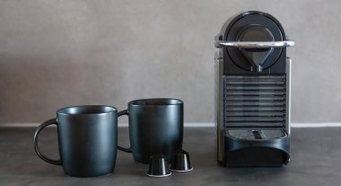 Kahve makinesi espresso ya da kapuçino yapıyor. Siyah renk makinesi, kapsüller ve kupalar mutfak masasında, ön manzara