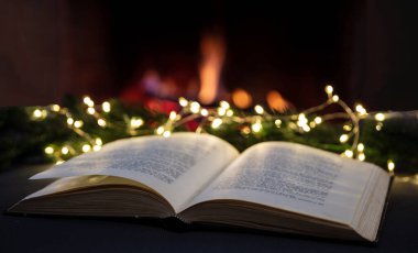 Noel okuması ve şöminenin yanında dinlenme, kitap açık, Xmas ağacı süsleme ışıkları, yanan arka plan bulanık şömine. Kış tatili sıcacık ev içi. 