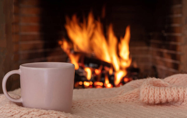 Cup of coffee on a sofa blanket, burning fireplace background. Pink color tea mug, relaxation by the fire, Christmas holiday cozy warm home