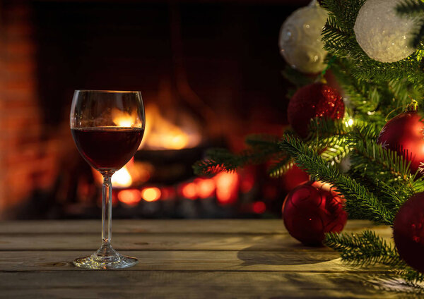  Christmas, red wine glass on a wooden table, burning fireplace background. Xmas tree decoration. Cozy warm home interior, winter holidays relaxation