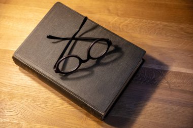 Masanın üzerinde kapalı eski siyah bir defter. Ahşap arka planda sert kapaklı defter ve gözlük, üst manzara. Öğretmenler masası.,