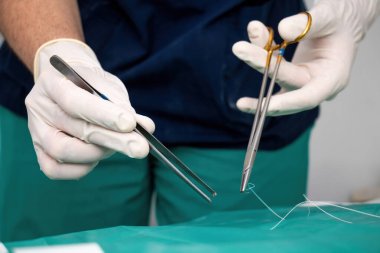 Elinde tek kullanımlık eldiven, elinde makas olan doktor cerrah yeşil kumaşın üzerine dikiş atarak yarayı ya da kesiği dikmiş. Ameliyat için paslanmaz araçlardan oluşan tıbbi ekipmanlar.