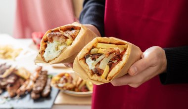 Gyro pita Shawarma. Yunan yemeği, dilimlenmiş et, patates, domates ve tzatziki, erkek eline sarılı kağıt, mutfak arka planı, yakın görüş.