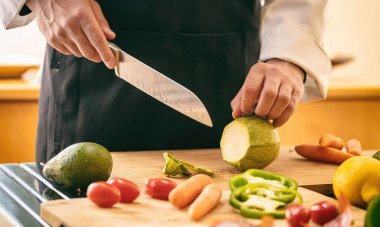 Siyah önlüklü bir şef kesim tahtasına kabak kesiyor, bıçağı yakın mesafeden tutuyor. Mutfak masasında taze sebze, restoranda vejetaryen yemeği..