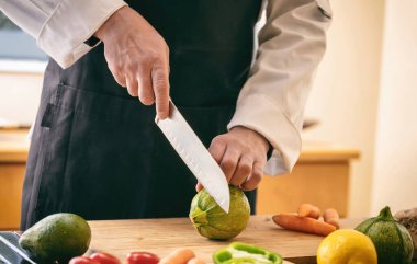 Siyah önlüklü bir şef kesim tahtasına kabak kesiyor, bıçağı yakın mesafeden tutuyor. Mutfak masasında taze sebze, restoranda vejetaryen yemeği..