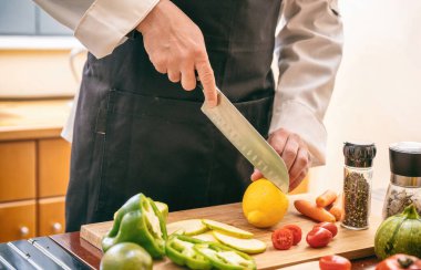 Siyah önlüklü bir şef doğrama tahtasına limon kesiyor, eli bıçak tutuyor. Mutfak masasında taze sebze, restoranda vejetaryen yemeği..