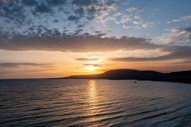 Günbatımında Yunan adası kıyısı. Dramatik gökyüzü, güneş batıyor, yelkenli silueti ve dalgalı deniz suyundaki yansımalar. Sakin ve huzurlu, yaz tatili dinlenmesi