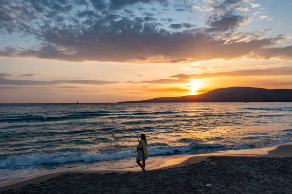 Denizin üzerinde gün batımı. Kumsalda yürüyen bir kadın. Islak kumda küçük dalgalar, renkli bulutlu gökyüzü arkaplanı. Elafonisos Yunan adası. Yunanistan.