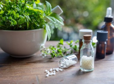 Homeopati eczanesi, bitkisel ilaçlar, doğal ilaçlar. Cam bir şişeden saçılan homeopatik küreler, havanda taze bitkiler, yeşil doğa arka planı..