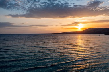 Günbatımında Yunan adası kıyısı. Dramatik gökyüzü, güneş batıyor, yelkenli silueti ve dalgalı deniz suyundaki yansımalar. Sakin ve huzurlu, yaz tatili dinlenmesi