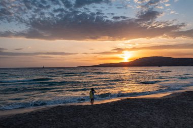Kumsalda gün batımını seyreden bir kadın. Islak kumda küçük dalgalar, renkli bulutlu gökyüzü arkaplanı. Elafonisos Yunan adası. Yunanistan.