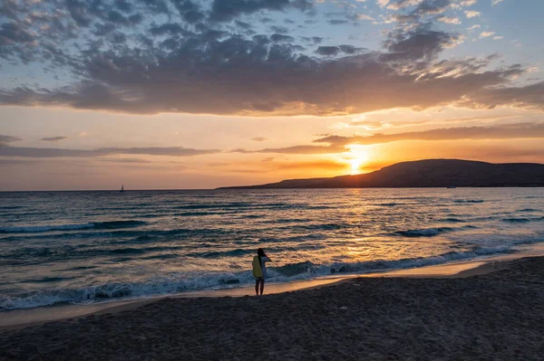 Kumsalda gün batımını seyreden bir kadın. Islak kumda küçük dalgalar, renkli bulutlu gökyüzü arkaplanı. Elafonisos Yunan adası. Yunanistan.