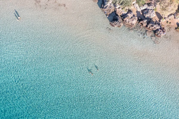 People swim in transparent turquoise blue sea water, people relaxing at the sandy beach or on the rocks, aerial drone view. Summer leisure under Greek sun.