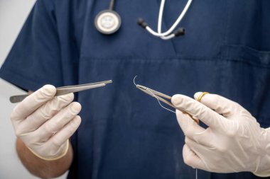 Tıbbi üniformalı doktor elinde tek kullanımlık eldiven, cerrahi alet, forseps, iğne tutacağı ve dikiş ipliği tutuyor. Boynunda steteskop olan bulanık bir doktor..