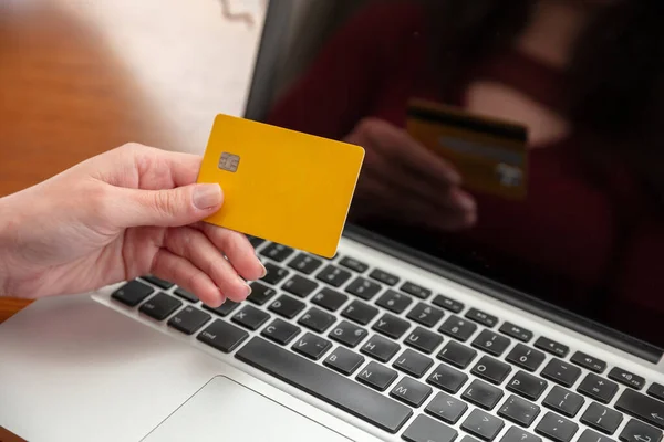Online shopping, credit card payment. Female hands holding blank yellow plastic card and using computer laptop, close up view, copy space.