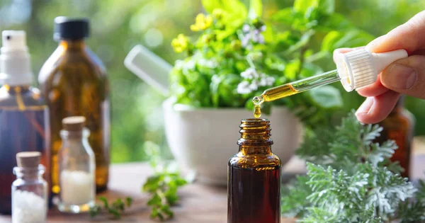 Alternative health medicine. Essential oil drop falling in a glass bottle, close up. Fresh herbs in a mortar on a wooden table, blur green nature background