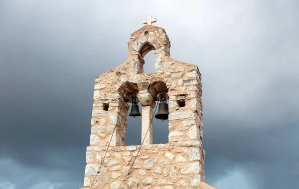 Belfry with two metal bronze bells at Mani Laconia, Peloponnese Greece. Under view of traditional stonewall bell tower with cross, cloudy sky background. Orthodox religious destination.