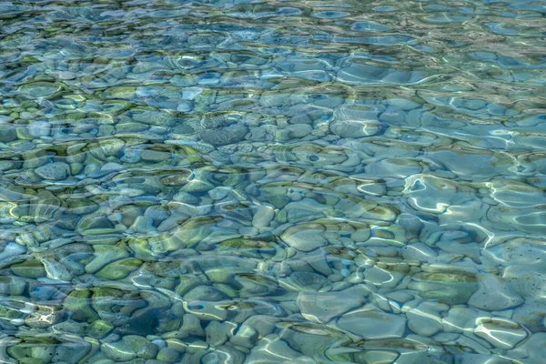 Clear crystal shallow blue sea water and rocky seabed background, texture. Calm transparent water surface, overhead view of underwater pebble, sun reflection.