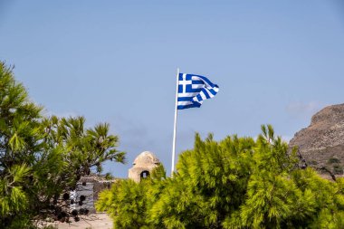 Greek flag waves in front of old building at Venetian Castle or Fortezza, monument at Kythira Ionian islands, Greece. View over green tree, blue sky background.