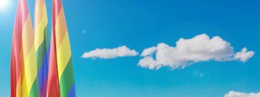 Pride flags on poles, blue cloudy sky background, banner. LGBT rainbow colors symbol, copy space. 3d render