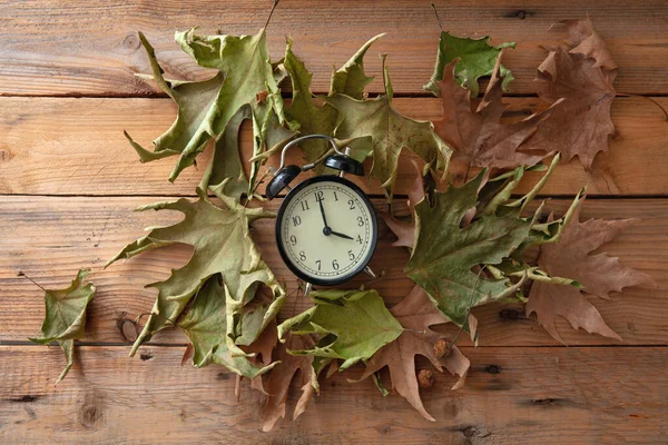 Daylight Saving Time, Fall Back. Black alarm clock and autumn leaves on wooden table, top vie