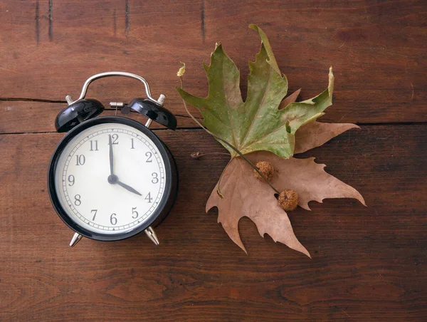 秋に戻る 昼光時間を節約 木製のテーブルの上に黒い目覚まし時計と秋の葉 トップビー — ストック写真