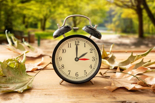 昼光時間を節約し 秋に戻る1時間 木製のテーブルの上の時間変化と黒の目覚まし時計 秋の木々と葉の背景 — ストック写真