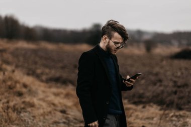 Siyah ceketli genç şık adam soğuk havada şehrin dışında akıllı telefon kullanıyor..