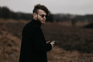 Siyah ceketli genç şık adam soğuk havada şehrin dışında akıllı telefon kullanıyor..