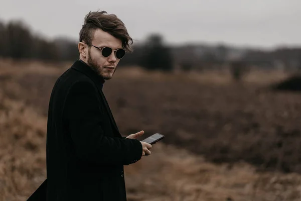 黒のコートを着た若いスタイリッシュな男が寒さの中都会の外でスマートフォンを使って — ストック写真