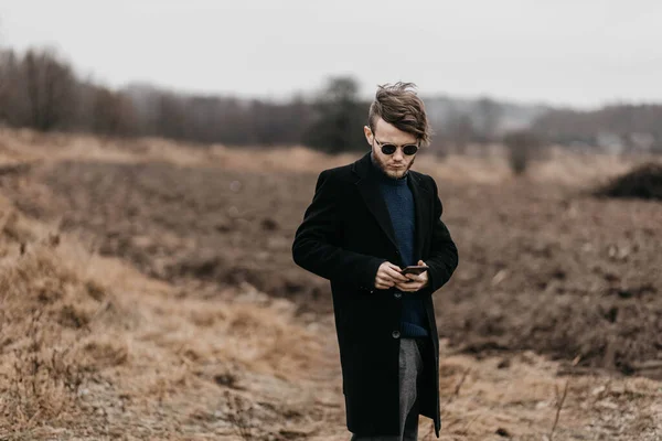 黒のコートを着た若いスタイリッシュな男が寒さの中都会の外でスマートフォンを使って — ストック写真