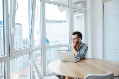 Gözlüklü, sakallı bir adam, pencerenin yanındaki masada dizüstü bilgisayarla çalışır ve düşünür. İnternet işi.