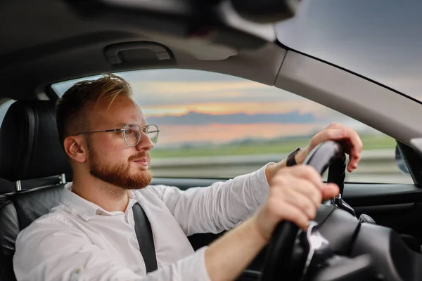 眼鏡やビジネス用の服を着た若い男性ドライバーが日没に乗っています ビジネスクラスのドライバーコンセプト ロイヤリティフリーのストック画像