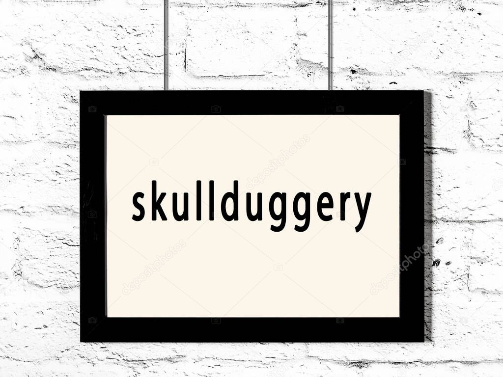 Black wooden frame with inscription skullduggery hanging on white brick wall 