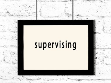 Black wooden frame with inscription supervising hanging on white brick wall 