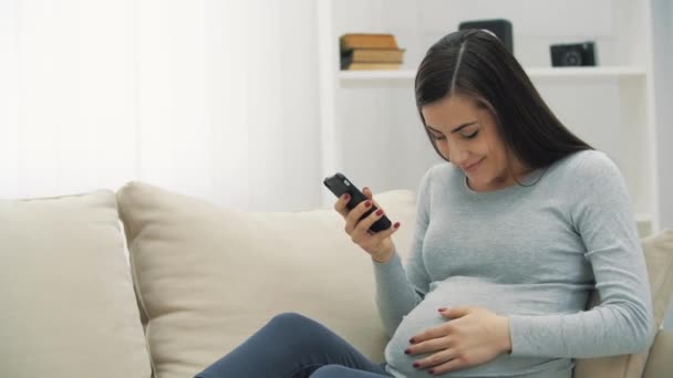 4k видео беременной женщины с телефоном на белом диване. — стоковое видео