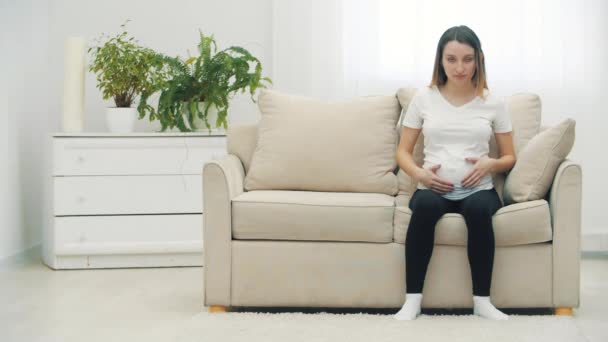 4k video de la mujer embarazada sentada en un sofá blanco y tocando su estómago. — Vídeo de stock
