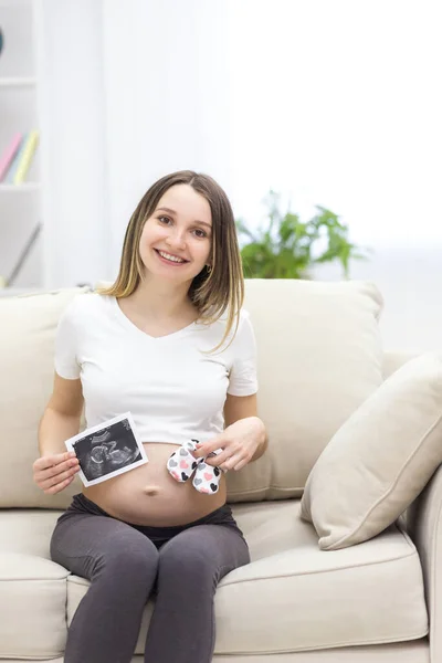 Foto einer schwangeren Frau, die auf dem Sofa sitzt und einen Ultraschallbericht hält. — Stockfoto