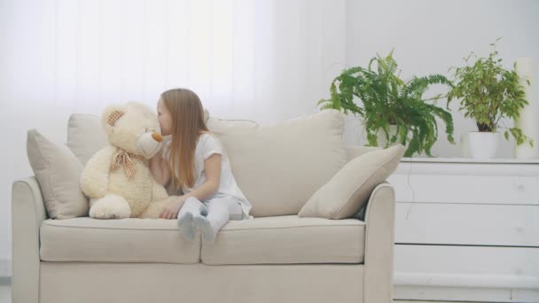 Vidéo ralenti 4k de petite fille partageant des secrets avec un ours en peluche sur le canapé. — Video