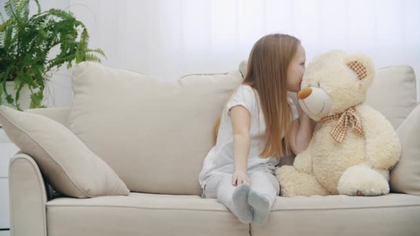 Vidéo 4k de petite fille partageant des secrets avec un ours en peluche sur le canapé. — Video