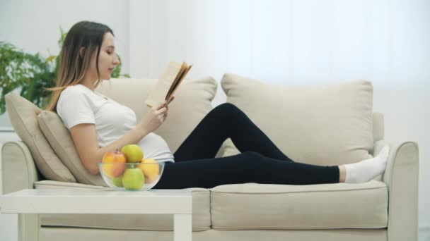 4k βίντεο με έγκυο γυναίκα που διαβάζει ένα βιβλίο και τρώει ένα μήλο. — Αρχείο Βίντεο
