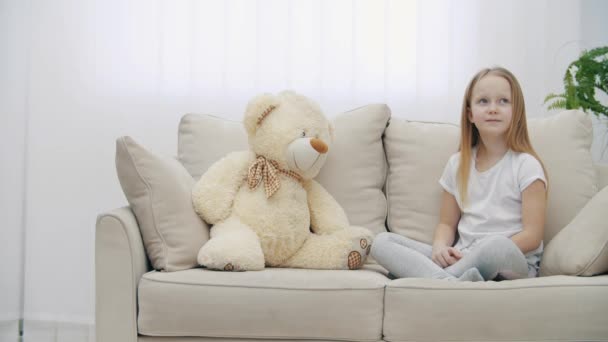 4k slow motion vídeo of little girl and teddy bear sitting on white sofa. — Vídeo de Stock