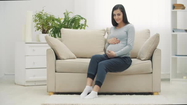 4k vídeo de mulher grávida sentada no sofá branco e tocando seu estômago. — Vídeo de Stock