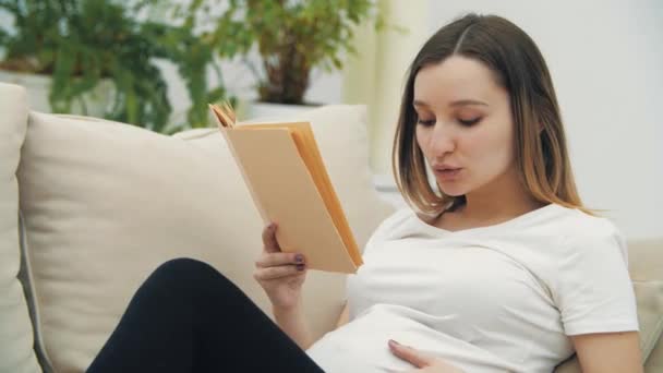 4k видео беременной женщины на диване, читающей книгу. — стоковое видео
