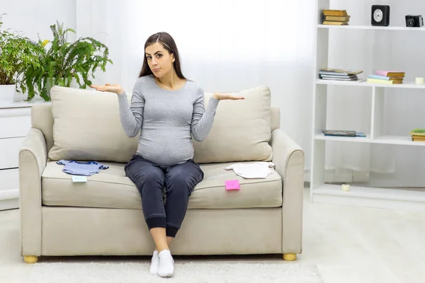Fotografie těhotné ženy sedící na pohovce s dětským oblečením. — Stock fotografie