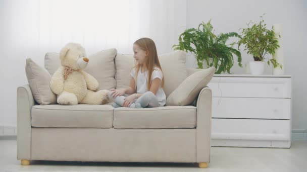 4k вялое видео маленькой девочки в белой одежде, играющей на белом диване с плюшевым медведем. — стоковое видео