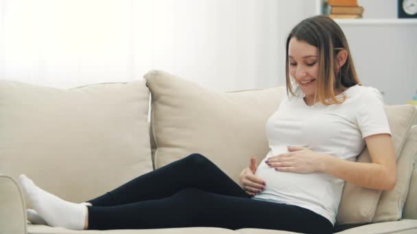 4kビデオの妊娠中の女性座っていますのホワイトソファとタッチ彼女の胃. — ストック動画