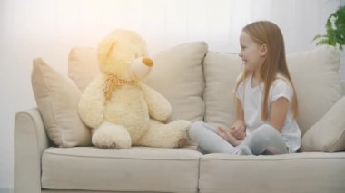 Beyaz elbiseli küçük bir kızın beyaz koltukta oyuncak ayıyla oynadığı 4K yavaş çekim videosu..